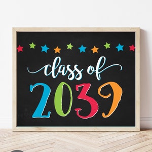 May include: A framed chalkboard sign with the words "class of 2039" in colorful lettering. The sign has a black background with a row of colorful stars across the top. The sign is in a light wood frame.