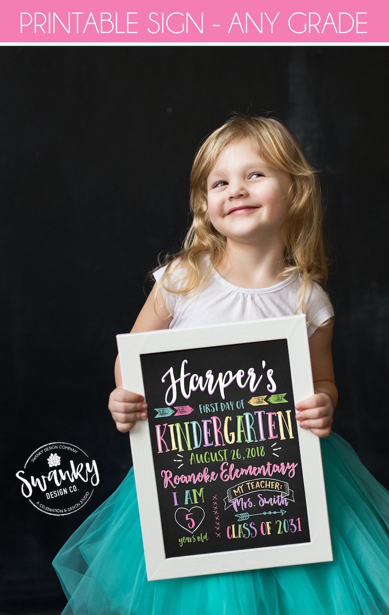 May include: A young girl smiles while holding a framed chalkboard sign. The sign is black with colorful text that reads "Harper's First Day of Kindergarten" and includes the date, school name, and teacher's name.