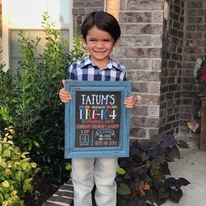 May include: A young child holding a blue framed chalkboard sign. The sign reads "Tatum's First Day of Pre-K 4" with the date September 6, 2017, and the name of the school. The child is wearing a plaid shirt and light-colored pants.