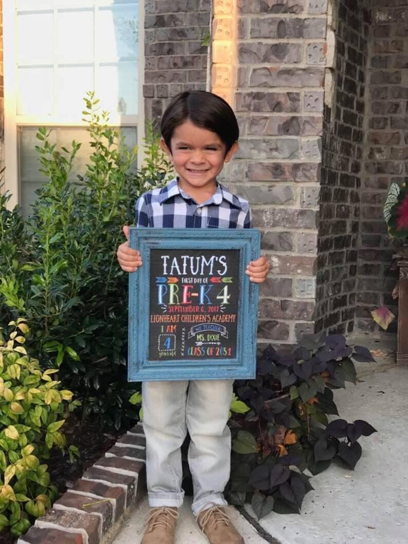 May include: A young child holding a blue framed chalkboard sign. The sign reads "Tatum's First Day of Pre-K 4" with the date September 6, 2017, and the name of the school. The child is wearing a plaid shirt and light-colored pants.