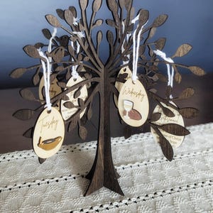 May include: A dark brown wooden tree with laser-cut leaves and branches. Oval-shaped wooden tags hang from the branches with white string. The tags are labeled with days of the week and small illustrations. The tree sits on a white patterned surface.