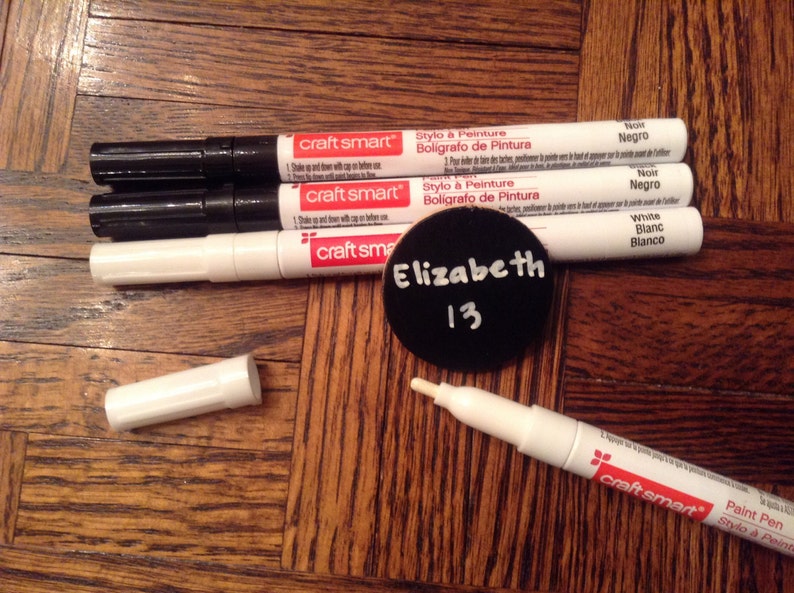 May include: Four white paint markers with black caps and red labels that say "craft smart" and "Stylo &agrave; Peinture Boligrafo de Pintura". The markers are labeled "Noir Negro", "Noir Negro", and "White Blanc Blanco". A white paint marker with a black cap and red label that says "craft smart" and "Paint Pen Stylo &agrave; Peinture". A black circle with white text that says "Elizabeth 13".