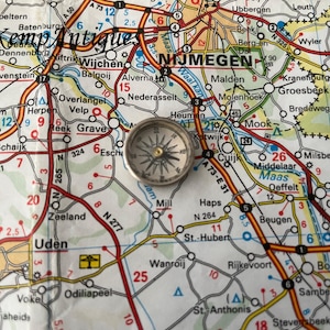 May include: A vintage compass with a silver case and a black face. The compass is on top of a map of the Netherlands, with the city of Nijmegen highlighted.