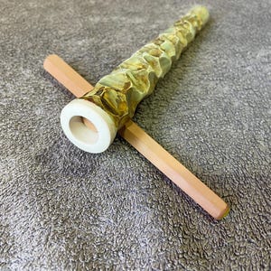Könnte beinhalten: Eine handgefertigte Holzpfeife mit einem hellbraunen Stiel und einem strukturierten, grün-goldenen Kopf. Die Pfeife liegt auf einer hellbraunen Oberfläche und zeigt ihr einzigartiges Design und ihre Handwerkskunst.