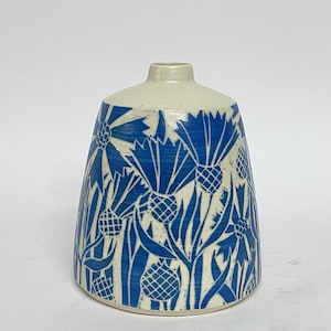 Puede incluir: Un jarrón de cerámica blanco con un patrón floral azul. El jarrón tiene una base ancha y un cuello estrecho. El patrón floral presenta flores y hojas azules estilizadas.