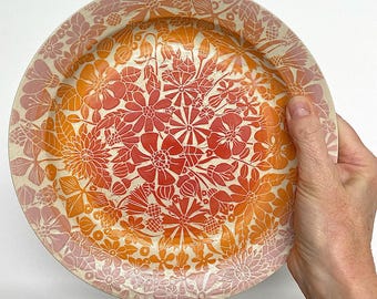 May include: A round ceramic plate with a floral design. The plate features a cream-colored base with a pattern of red, orange, and pink flowers. The flowers are arranged in a dense, repeating pattern across the surface of the plate.
