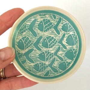 Könnte beinhalten: Eine kleine, runde Keramikschale mit einem blaugrünen Glazure. Die Schale ist mit einem weißen Design von Fischen verziert, die in Wellen schwimmen.
