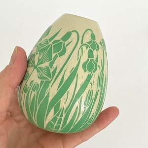 Könnte beinhalten: Eine weiße Keramikvase mit grünem Blumendesign. Die Vase hat die Form eines Eies und eine glatte, glänzende Oberfläche.