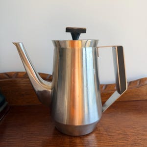 May include: A stainless steel coffee pot with a tapered body, a long angled spout, and a flat-topped black handle. The lid features a small black knob handle at the centre. The metal surface has a brushed finish with subtle vertical reflections.