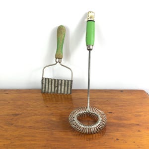 May include: Two vintage kitchen utensils on a wooden surface. One is a crinkle cutter with a green handle. The other is a wire whisk with a green handle and a decorative top. Both tools have a well-worn, antique appearance.