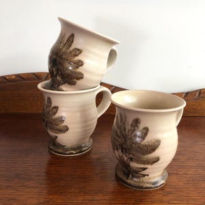 May include: Four beige ceramic mugs with a brown floral design. The mugs have a rounded shape with a handle and a flared rim. The mugs are stacked and arranged on a wooden surface.