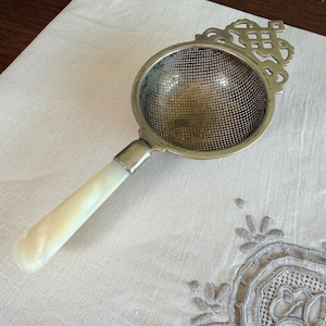 May include: A vintage tea strainer with a silver mesh bowl and a decorative handle. The handle is made of a light-coloured, possibly mother-of-pearl material. The strainer has an ornate, openwork design at the top.