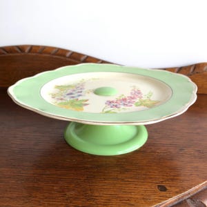 May include: A vintage cake stand with a scalloped edge. The stand has a light green base and rim, with a floral design in the centre. The floral design includes purple, blue, and yellow flowers. The stand has a gold trim.