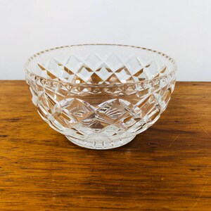 May include: A clear, diamond-cut glass bowl with a round shape. The bowl has a decorative pattern and sits on a small, circular base. The bowl is on a wooden surface.