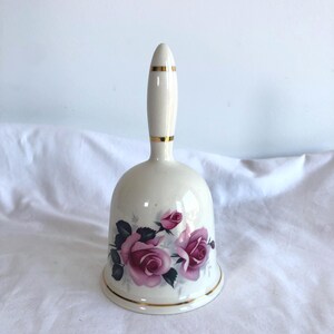 May include: A cream-coloured ceramic bell featuring a floral design of pink roses and green leaves. The bell has gold detailing and a handle. A decorative item for the home.