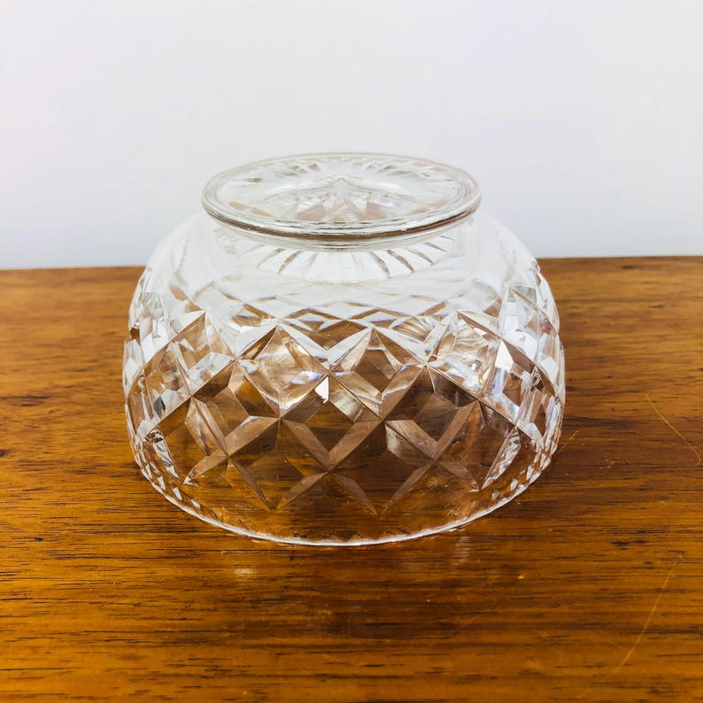 May include: A clear, diamond-cut glass bowl with a rounded shape. The bowl has a thick base and a decorative pattern of intersecting lines, creating a geometric design. The bowl is sitting on a wooden surface.