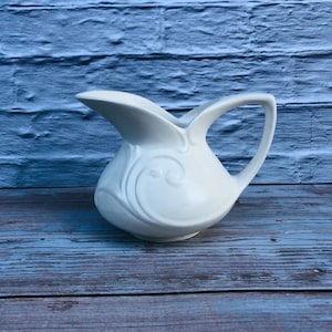 May include: A white ceramic jug with a curved handle and a spout. The jug features an embossed swirl design on the front. The jug is on a blue wooden surface.