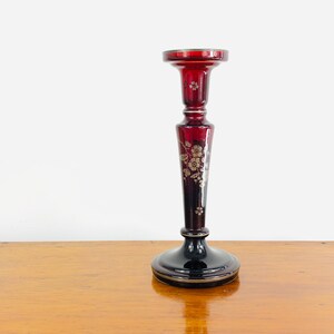 May include: A tall, ruby red glass candlestick with a black base and gold trim. The candlestick features a floral design in gold and silver. The candlestick is approximately 25 cm tall.