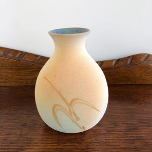 May include: A small, beige ceramic vase with a light blue rim and a rounded body. The vase has a simple, abstract brown design. The vase is displayed on a dark wooden surface.