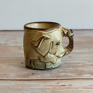 May include: A ceramic mug with a speckled beige and brown finish featuring a moulded elephant design. The elephant's trunk forms the handle of the mug, while the body of the elephant is embossed on the side of the vessel with visible eyes and ears.