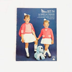 May include: A vintage knitting pattern booklet for babies and toddlers. The cover features two young girls wearing pink and white striped sweaters and skirts, holding a blue toy dog. The booklet is titled "Patons Knitting Book No. 714" and includes instructions for making twin sets, sweaters, caps, mitts, and socks.