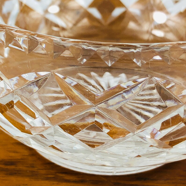 May include: Close-up of a clear, cut-glass bowl with a diamond pattern. The bowl's rim curves gently, and the light reflects off the facets, creating a sparkling effect. The background is a warm, wooden surface.