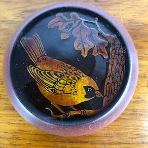 May include: A circular decorative item with a detailed illustration of a bird on a branch with leaves. The bird has yellow and brown feathers, set against a dark background. The item has a brown rim.