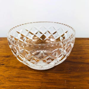 May include: A clear, cut-glass bowl with a diamond pattern. The bowl has a round shape with a textured rim and sits on a wooden surface. The bowl is empty and reflects light, showcasing its intricate design.