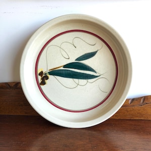 May include: Cream-coloured ceramic plate with a red border and a hand-painted design of green leaves and brown accents. The plate has a slightly raised edge. Diameter approximately 25 cm.