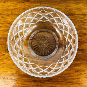 May include: Clear glass bowl with a diamond pattern around the rim and a circular design in the center. The bowl is sitting on a wooden surface.