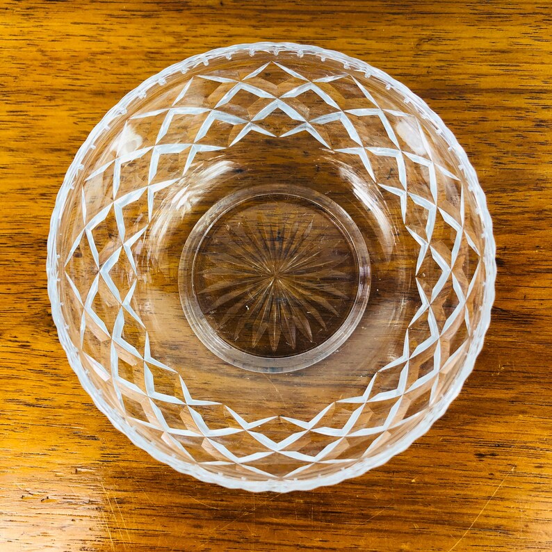 May include: Clear glass bowl with a diamond pattern around the rim and a circular design in the center. The bowl is sitting on a wooden surface.