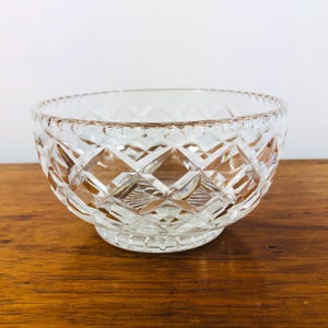 May include: A clear, cut-glass bowl with a diamond pattern. The bowl has a round shape with a slightly flared rim and a small, circular base. The bowl is sitting on a wooden surface.