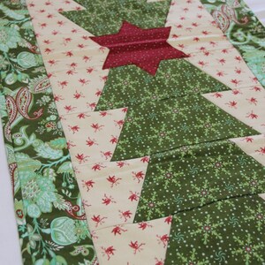 May include: A green, white, and red quilted table runner with a star design. The runner features a repeating pattern of green triangles with white snowflakes and a cream background with red birds. The runner is bordered with a green and white paisley pattern.
