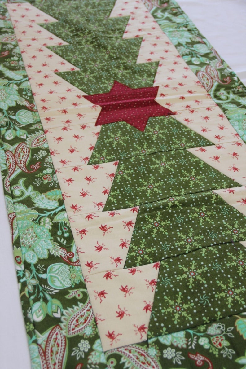 May include: A green, white and red quilted table runner with a star design. The runner features a repeating pattern of green triangles with white snowflakes and a cream background with red birds. The runner is bordered with a green and white paisley pattern.
