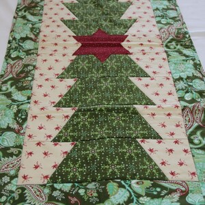 May include: A green and cream quilted table runner featuring a Christmas tree design. The tree is made from green fabric with a snowflake pattern and a red star at the top. The runner has a paisley border.