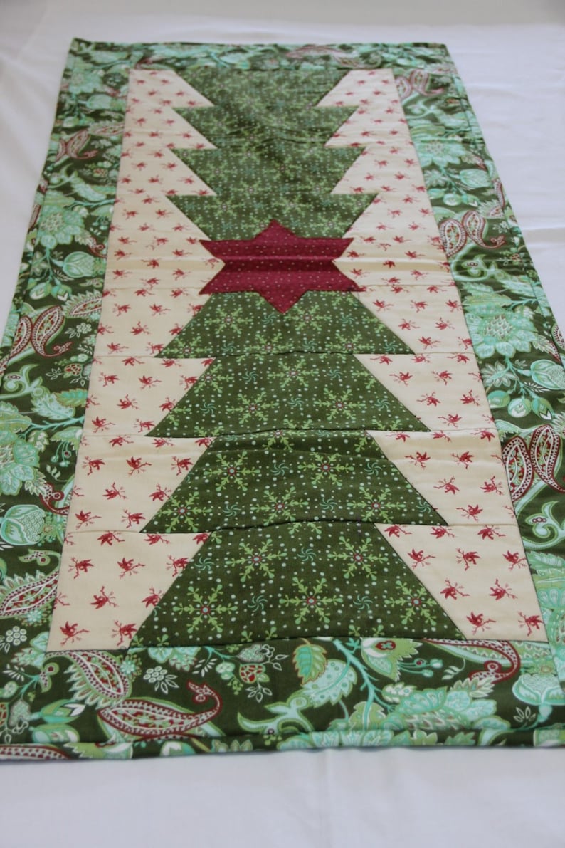 May include: A green and cream quilted table runner featuring a Christmas tree design. The tree is made from green fabric with a snowflake pattern and a red star at the top. The runner has a paisley border.