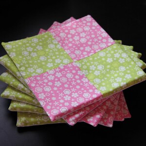 May include: A stack of six green and pink fabric coasters with a floral pattern. The coasters are made of cotton fabric and are ideal for protecting surfaces from heat and spills.
