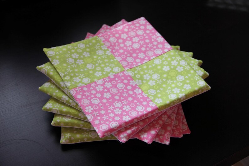 May include: A stack of six green and pink fabric coasters with a floral pattern. The coasters are made of cotton fabric and are ideal for protecting surfaces from heat and spills.