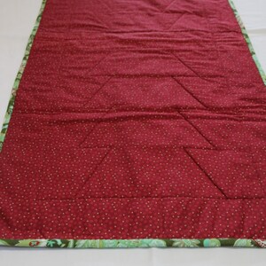 May include: A red quilted table runner with a green and white floral border. The runner has a diamond pattern quilted into the fabric.