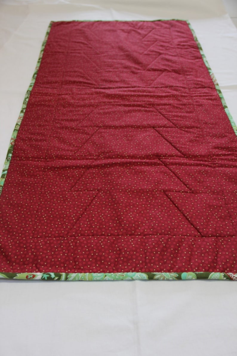 May include: A red quilted table runner with a green and white floral border. The runner has a diamond pattern quilted into the fabric.