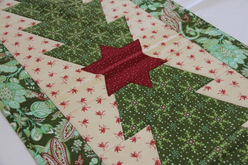 May include: A green and cream quilted table runner with a red star in the centre. The runner features a repeating pattern of snowflakes and small red flowers.