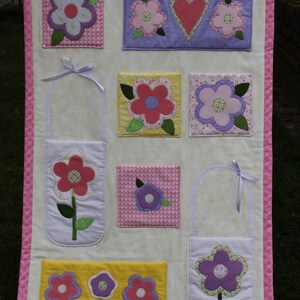 May include: A white fabric wall hanging with pink trim and multiple pockets. Each pocket features a different flower design with pink, purple, and yellow fabrics. The pockets are ideal for storing small items.