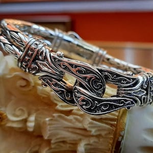 May include: A silver bracelet with a detailed, intricate design. The bracelet is made of two braided strands of metal that are connected by a large, ornate clasp.