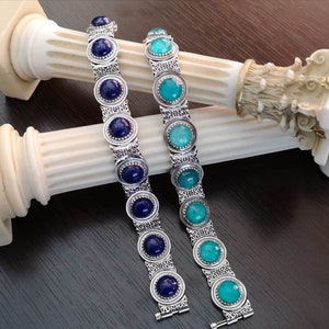 May include: Two silver bracelets with blue and turquoise gemstones. The bracelets have intricate silver designs and are arranged on a white column.