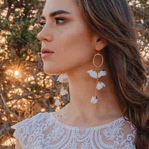 Könnte beinhalten: Eine Frau in einem weißen Spitzenkleid mit Blumenmuster. Sie trägt lange, baumelnde Ohrringe mit weißen Blüten und goldenen Akzenten. Das Kleid hat einen runden Ausschnitt und kurze Ärmel.