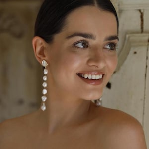 May include: A woman wearing a white off-the-shoulder dress with a pearl and gold drop earring.