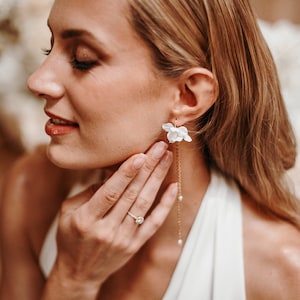 May include: A woman wearing a white dress with a delicate floral earring featuring a white flower with a gold chain and pearl accents.