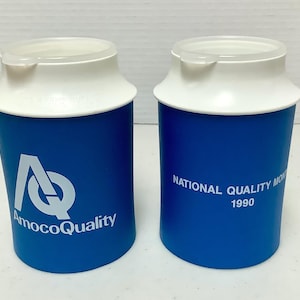 May include: Two blue and white travel mugs. One mug has the text "AmocoQuality" and a logo. The other mug has the text "NATIONAL QUALITY MONTH 1990". The mugs have a white lid with a small spout.