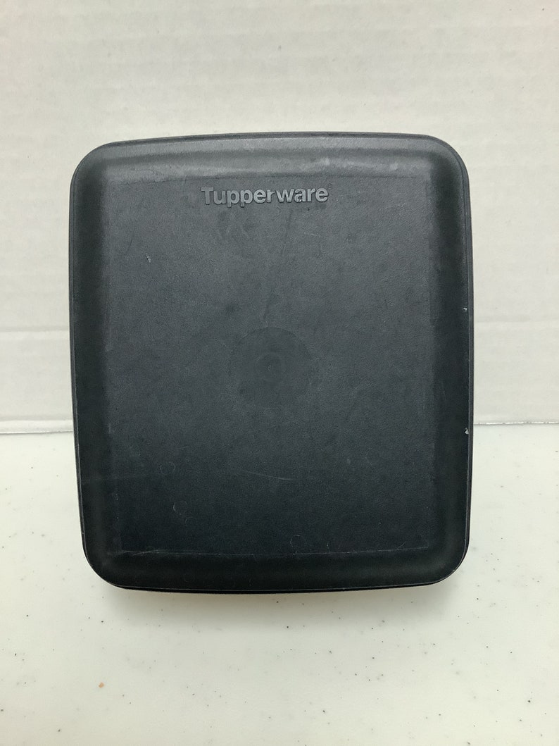 Puede incluir: Un recipiente Tupperware negro, cuadrado, con esquinas redondeadas. La palabra "Tupperware" est&aacute; grabada en la parte superior. El recipiente est&aacute; hecho de un material pl&aacute;stico duradero y parece estar en buenas condiciones.