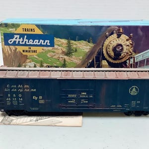 May include: Athearn Trains in Miniature model train car in a box. The box has a mountain and train scene. The model train car is dark blue with text and logos. The box reads "Athearn in Miniature".